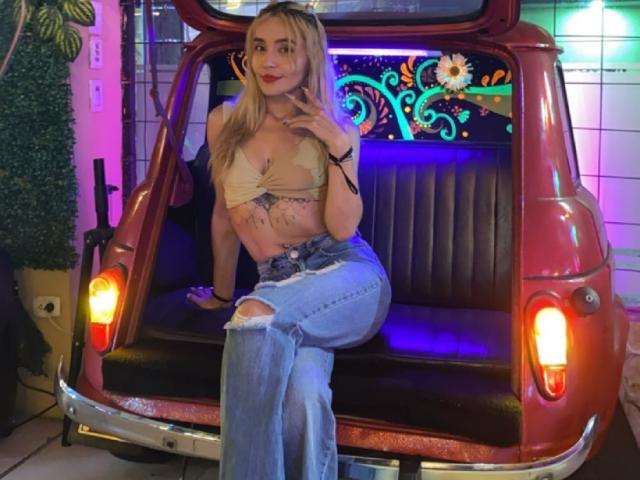 MindyLove with blonde hair, tan bra, and ripped jeans, sitting in a red car's open back, hand on chest, showing her tits and a tattoo on her stomach.
