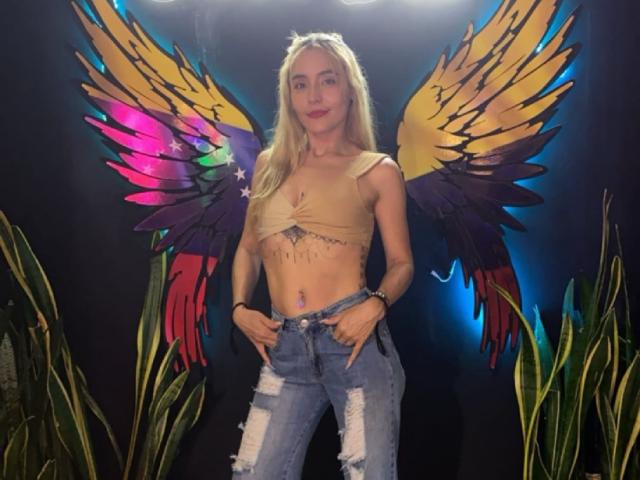 MindyLove with blonde hair, small tits, wearing a tan crop top and ripped jeans, standing in front of colorful, painted angel wings. Hands in jeans pockets, smiling.