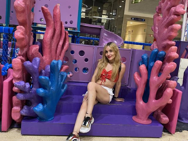 MindyLove, blond, in een rood bikini topje, witte shorts, zittend op een paarse bank, omringd door roze en blauwe structuren die op koraal lijken, lachend, met witte en zwarte sneakers, in een winkelcentrum..