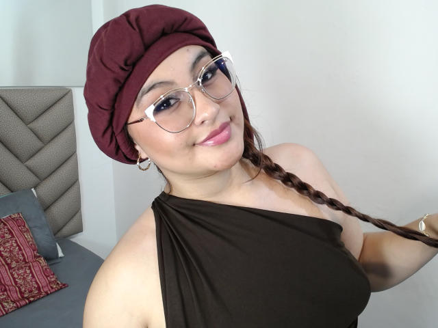 HelueJamil with glasses, maroon headscarf, brown braid, smiling, black sleeveless top, showing her tits, silver earrings, in a bedroom.