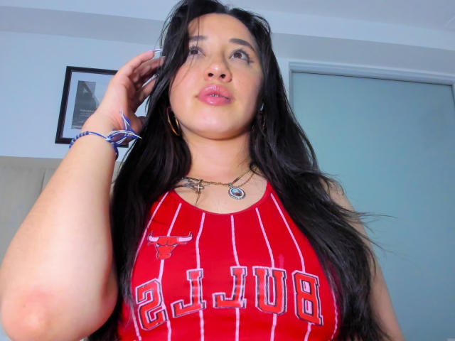 JessyBarnnett with long black hair, wearing a red tank top with white stripes and "CLUB" text, showing her big boobs, standing, hand in hair, looking up.