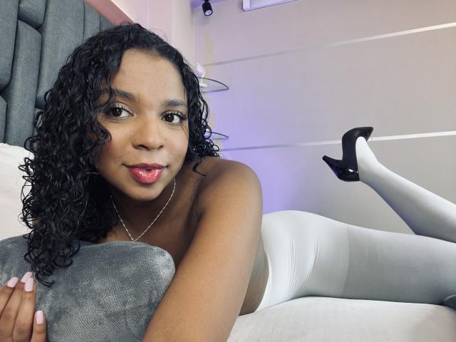 LisaMendez with curly black hair, medium brown skin, pink lipstick, lying on a bed, white leggings, black heels, holding a gray pillow, smiling at the camera, topless.