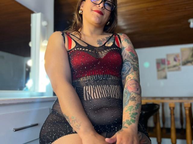 ValeriaCuttest with tattoos on her arm and chest, wearing a see-through black dress with red bra, showing her tits and pussy, sitting in a room with wooden ceiling.