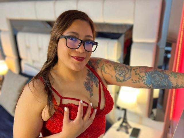ValeriaCuttest with glasses, red lace bra, and tattoos on her arm, holding her tits, smiling, in a bedroom with a tufted headboard and lamps.