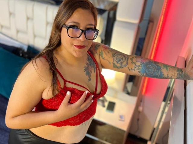 ValeriaCuttest with glasses, red lace bra, and tattoos, covering her tits with one hand, smiling, arm against red-lit wall, brown hair, red lipstick, black-framed glasses, and black pants.