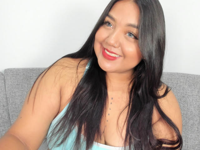 SexyChubbyHot with long black hair, blue eyes, red lipstick, and a smile, wearing a light blue tank top, showing small tits and a chest tattoo.