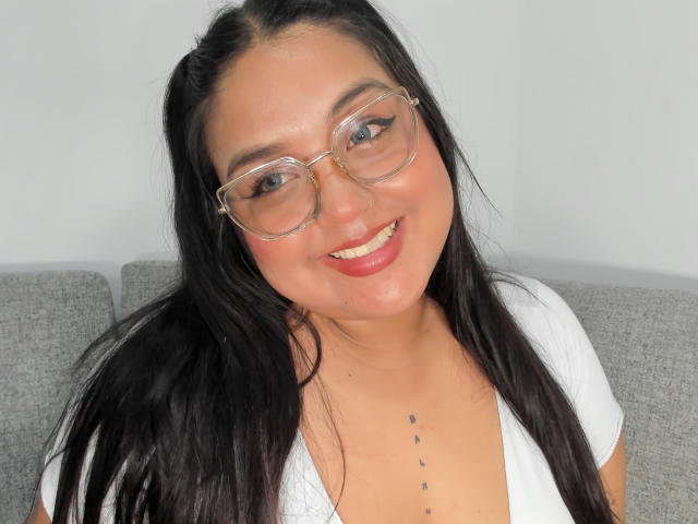 SexyChubbyHot with long black hair, clear glasses, white top, smiling, red lipstick, small black dots on chest, sitting on gray couch, looking at camera.