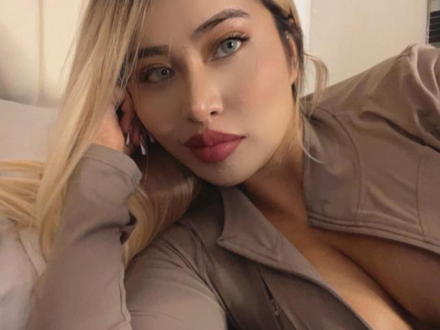 DahliaVelvet, con i capelli biondi, gli occhi azzurri e il rossetto rosso, indossa una camicia marrone che mette in mostra il décolleté, è sdraiata con una mano sul viso. Il seno è visibile..