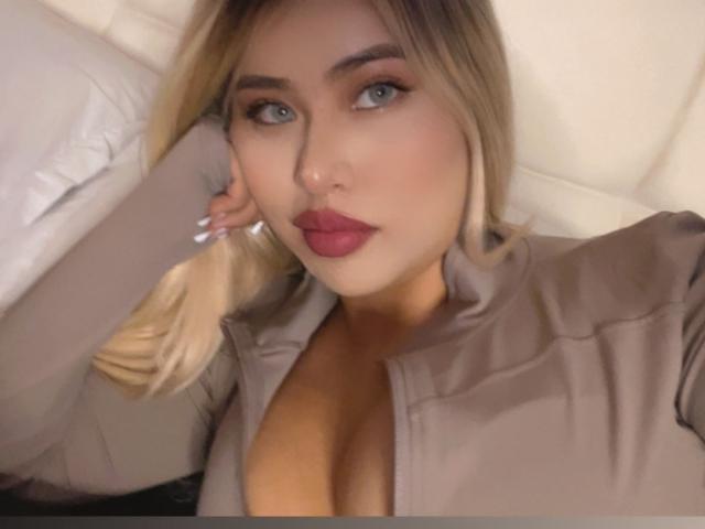 DahliaVelvet mit blonden Haaren, blauen Augen und rotem Lippenstift, trägt ein beiges Hemd, zeigt ihre Brüste, liegt auf einem weißen Kissen und schaut in die Kamera..