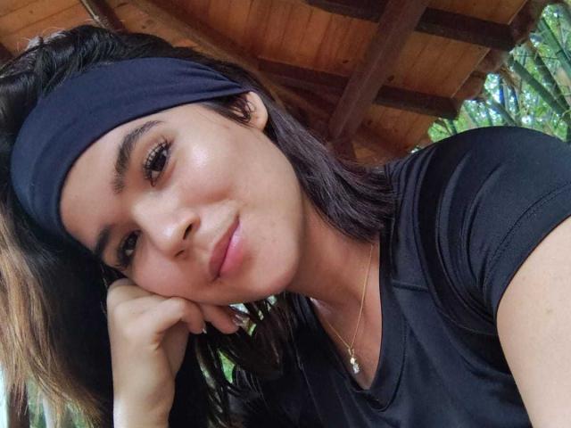 ScarlletZhen with black headband, brown eyes, and subtle smile, leaning on hand, wearing black shirt, necklace, and small pendant, with slightly shiny skin.