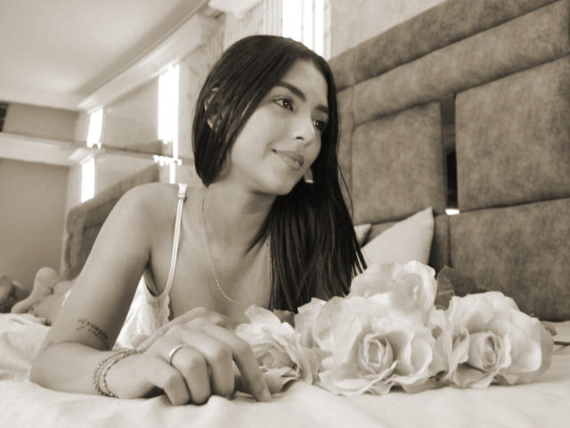 LunaBellas with long dark hair, lying on a bed, smiling, wearing a white dress, holding rose petals, visible tattoo on arm, ring on finger, sepia-toned.