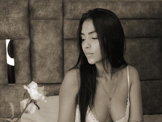 LunaBellas with long dark hair, wearing a low-cut white lace top, looking down, showing her tits, necklace with heart pendant, sitting in bed, flower in left.