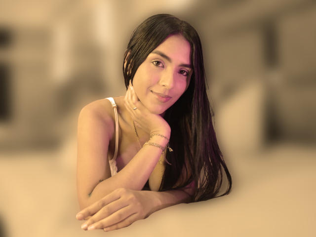 LunaBellas with long black hair, tan skin, small tits, wearing a white tank top, leaning on a surface, smiling softly, right hand touching her chin, blurred background.