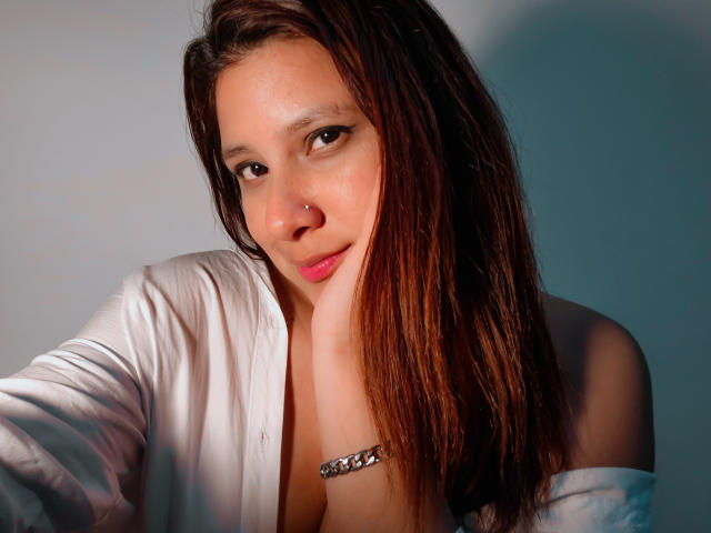 LadyStarOne, com cabelo castanho, piercing no nariz e lábios rosados, vestindo uma camisa branca, tem a mão no rosto, mostrando os seios, e usa uma pulseira de prata..