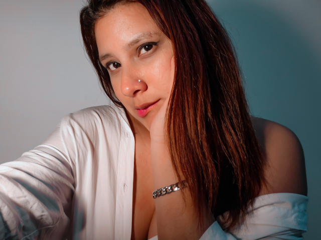 LadyStarOne, avec ses cheveux bruns mouillés, une chemise blanche, tombant d'une épaule, dévoilant sa poitrine, un piercing au nez et un bracelet en argent, regardant la caméra, avec un léger sourire, une lumière douce sur son visage..