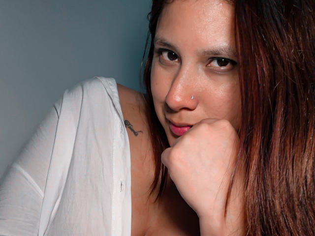 LadyStarOne, com olhos castanhos, piercing no nariz e cabelo castanho molhado, vestindo uma camisa branca, com o queixo apoiado na mão, mostrando uma tatuagem no ombro esquerdo..