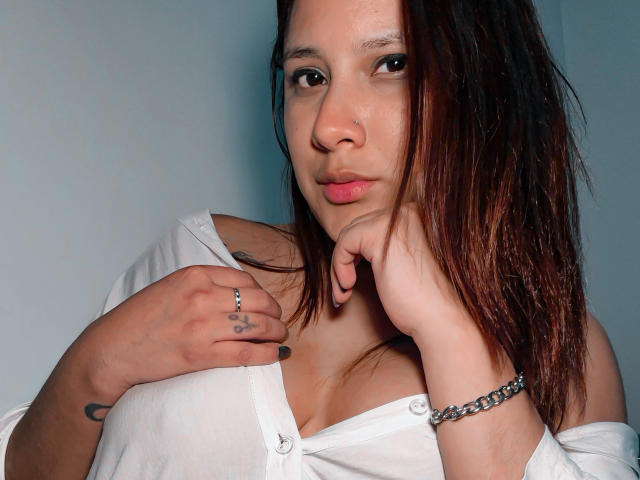 LadyStarOne with brown hair, white shirt, and silver bracelet, showing off her tits with one hand on her chest and the other on her neck. Tattoo on left hand.