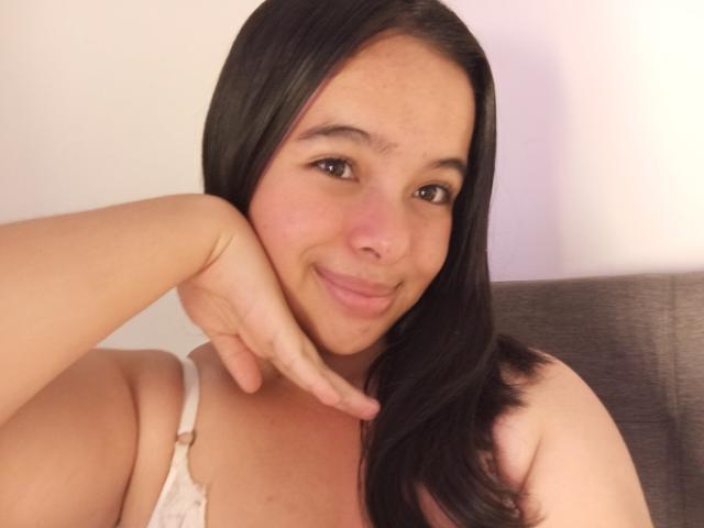 SolMartinez has black hair, a light skin tone, and is smiling at the camera with her hand under her chin, wearing a white bra, showing her tits.