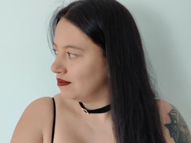 IsabelaDeLuca has black hair, red lipstick, and a tattoo on her right arm. She wears a black choker and thin-strapped top, looking to the side.