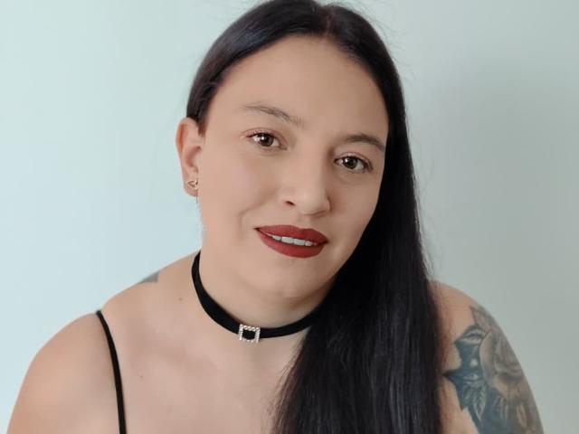 IsabelaDeLuca has long black hair, red lipstick, and a black choker with a silver buckle, showing a tattoo on her right arm, and small ear studs.