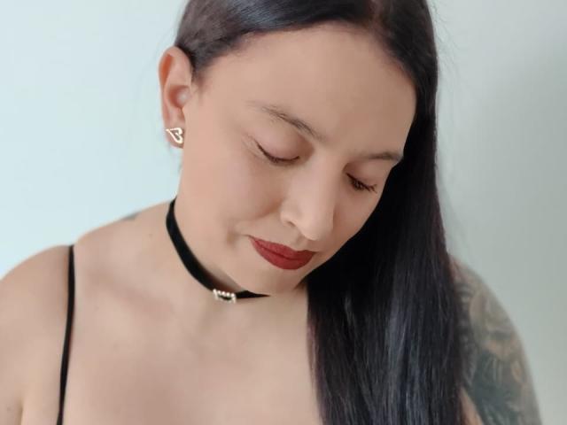 IsabelaDeLuca with closed eyes, black choker, red lipstick, straight black hair, small triangular earrings, black spaghetti strap top, visible tattoo on right shoulder, pale skin, slight smile.