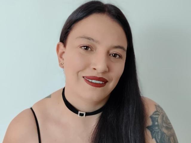 IsabelaDeLuca has long black hair, red lipstick, a black choker with a silver buckle, and visible tattoos on her right shoulder. She's smiling with brown eyes.