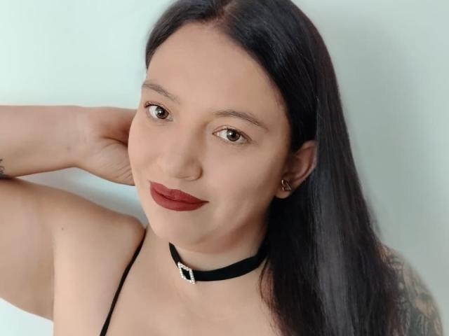 IsabelaDeLuca with dark hair, red lipstick, black choker, and small tits, smiling, hand behind head, tattoo on arm, ear piercing, soft light, light skin, brown eyes.