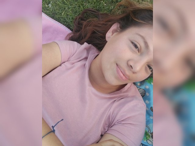 ScarlettAmelia lying on grass, pink t-shirt, brown hair, smiling, soft light, blurry out-of-focus face on right, pink blanket, relaxed, casual, natural, slight tan, cute.