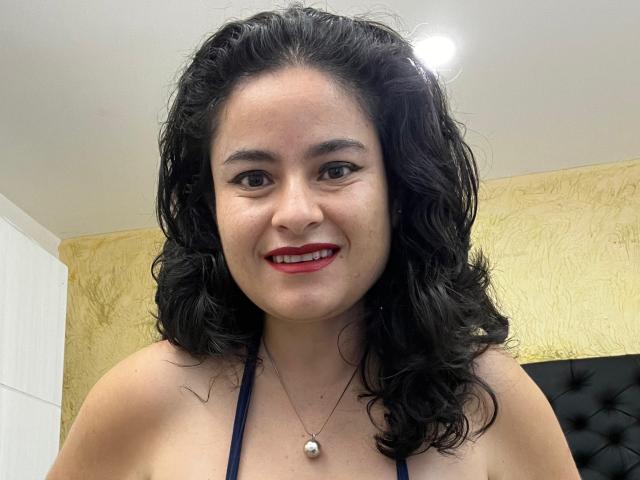 Sara, avec ses cheveux noirs bouclés, son rouge à lèvres rouge et son collier de perles, sourit à l'appareil photo, montrant ses épaules nues et un haut noir à encolure américaine. Ses seins sont visibles..