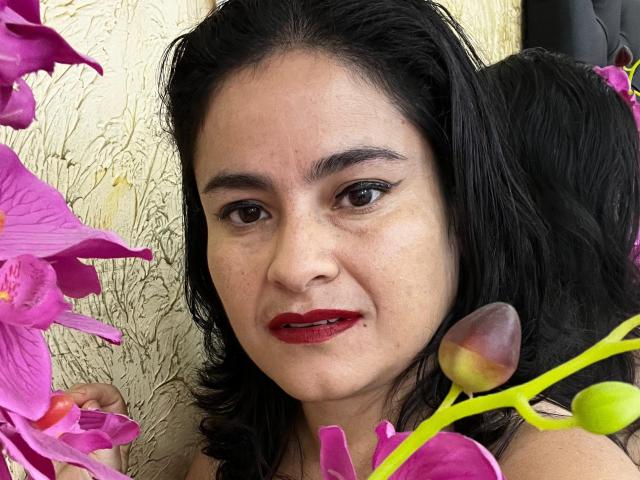 SaraBrendas hat dunkles Haar, roten Lippenstift und braune Augen, im Vordergrund befinden sich rosa Orchideen. Ihr Hintern ist nicht sichtbar. Sie trägt einen neutralen Gesichtsausdruck..