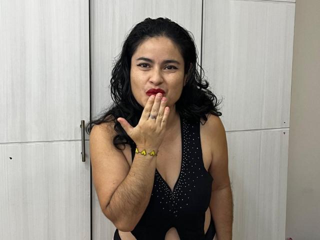SaraBrendas, com cabelos cacheados escuros, vestido preto de alça e pulseira amarela, chupa o dedo com batom vermelho. Sua mão cobre a boca. Ela está em um quarto..