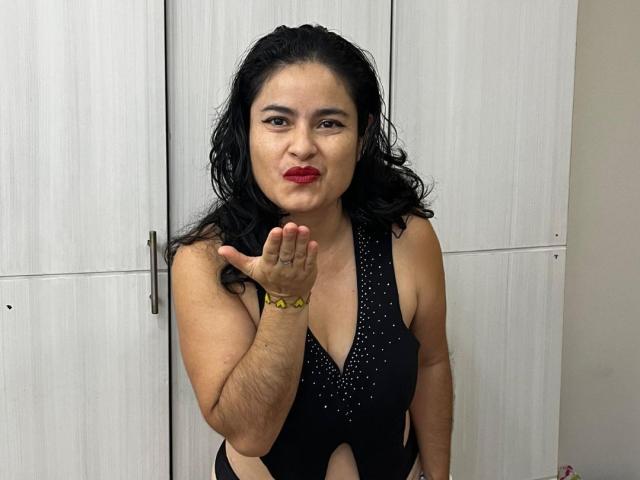 Sara, con i capelli neri e ondulati, rossetto rosso e costume da bagno nero con borchie, sta mandando un bacio, mostrando la sua mano con un braccialetto giallo e il suo seno di media grandezza..