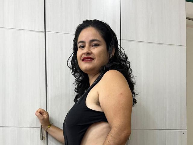Sara, mit lockigem, schwarzem Haar, rotem Lippenstift und einem schwarzen Tanktop, zeigt den linken Achselbereich und kleine Brüste, steht lächelnd vor weißen Schränken..