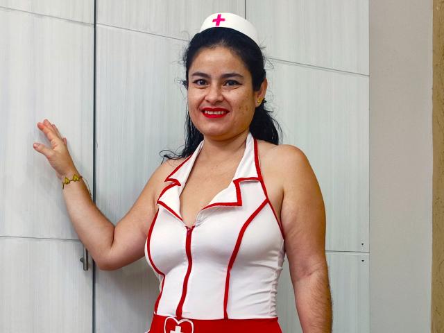 SaraBrendas, mulher asiática, vestindo um uniforme de enfermeira com detalhes vermelhos, touca branca, sorrindo, batom vermelho, cabelo preto em um rabo de cavalo, mão direita levantada, parede de madeira branca ao fundo..