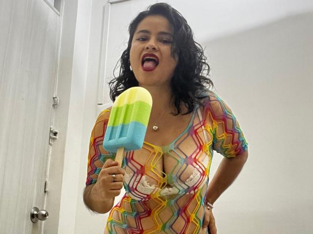 Sara, avec ses cheveux noirs bouclés, la langue tirée, portant une robe colorée à motifs en zigzag, dévoilant ses seins avec des pasties, tenant un popsicle arc-en-ciel, se tient devant une porte blanche..