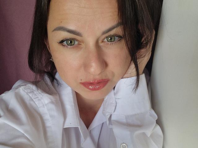 JoyArinka with green eyes, glossy pink lips, white shirt, and dark brown hair, looking directly at the camera with a slightly serious expression. She has a small mole on her chin.