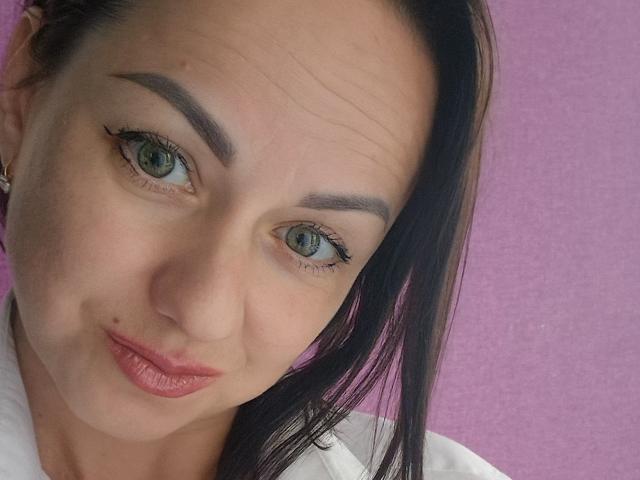 JoyArinka has blue eyes, dark eyebrows, and pink lips. Her black hair is straight, and she's wearing a white shirt. Her face has a small mole on the right side.