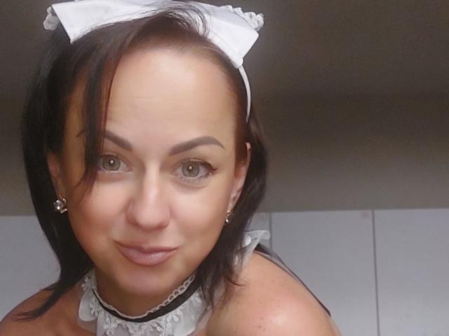 JoyArinka with medium brown skin, green eyes, black hair, white maid headband, lace choker, and earrings, smiling subtly, showing her tits slightly with a cute, sexy look.
