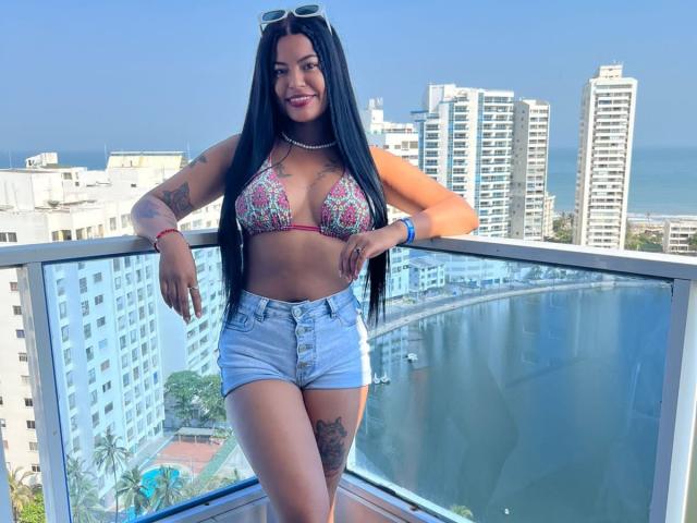 DannaHarmonyy with long black hair, wearing a colorful bra, blue shorts, tattoos, and sunglasses on a balcony, smiling at the camera, showing her tits and ass.