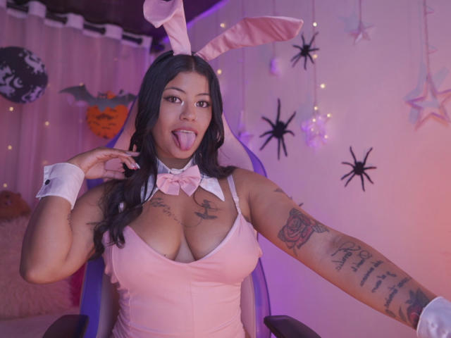 DannaHarmonyy in a pink bunny costume with large tits, tattoos on arms, sticking out tongue, wearing pink bowtie, white cuffs, and pink bunny ears, sitting in chair.
