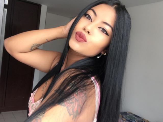 DannaHarmonyy with long black hair, pouty red lips, tattooed arm, and pink lace bra, posing with hand in hair, looking at camera, in a simple room.