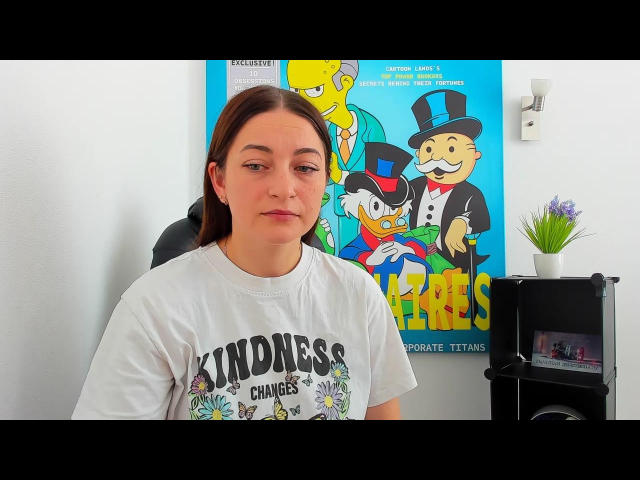 AmoraMiia with medium brown hair, white shirt saying "Kindness Changes," neutral expression, slight smirk, in front of colorful poster with animated characters. She has a cute ass.