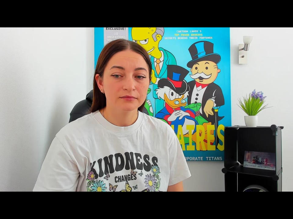 AmoraMiia with medium brown hair, white shirt saying "Kindness Changes," neutral expression, slight smirk, in front of colorful poster with animated characters. She has a cute ass.