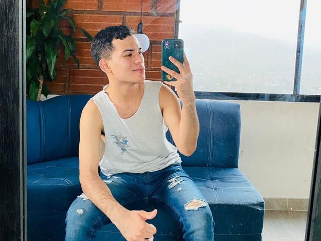 Alessandro Musk, con cabello negro corto, vistiendo una camiseta gris sin mangas y jeans azules rotos, se toma un selfie con un teléfono, sentado en un sofá azul, sonriendo ligeramente..