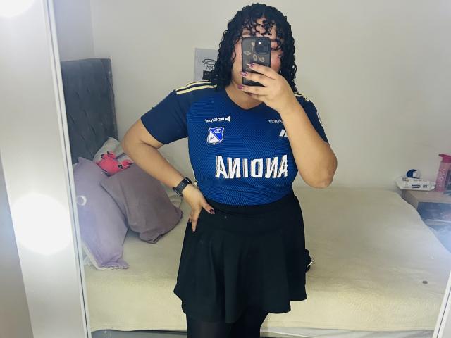 KarlaWild with curly black hair, wearing a blue Ammara sports jersey and black skirt, holding a phone in front of her face, standing in a mirror selfie.