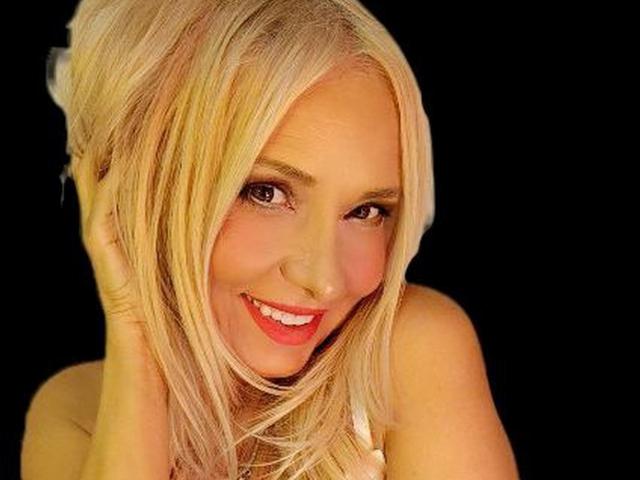 Leily with blonde hair, smiling with red lips, hand in hair, black background, showing off her tits, cute and sexy, smiling eyes, looking at camera.