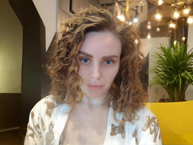 MiaGrantes a les cheveux bruns bouclés, les yeux bleus et une expression sérieuse. Elle porte une robe blanche à motifs floraux, qui laisse entrevoir sa poitrine. Des lumières vives au plafond et un pot jaune se trouvent en arrière-plan..