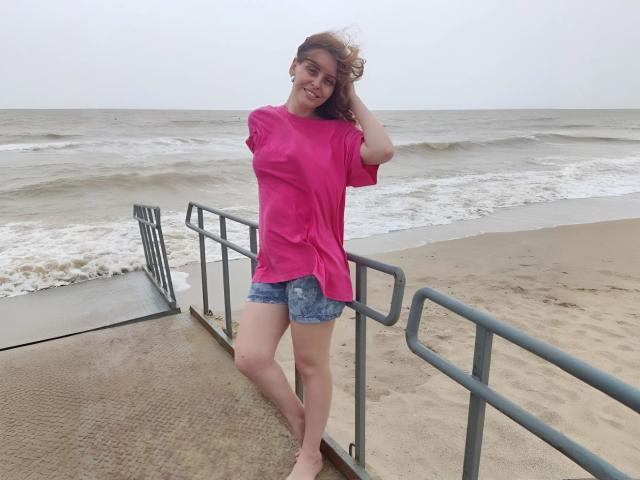 MiaGrantes stands barefoot on a sandy boardwalk, wearing a pink shirt and blue denim shorts, smiling with her hand in her brown hair, waves in the background.