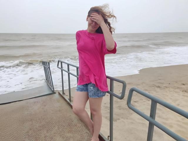 MiaGrantes, avec ses cheveux bruns ondulés, son t-shirt rose et son short en jean, se tient sur une rambarde en métal, les yeux cachés, souriante, sur une plage de sable avec de douces vagues..