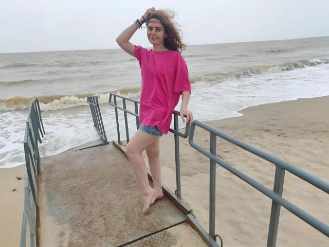MiaGrantes, zonder schoenen, in een roze shirt en denim shorts, staat op een metalen trap aan de kust, met één hand in haar krullende bruine haar, en glimlacht naar de camera..